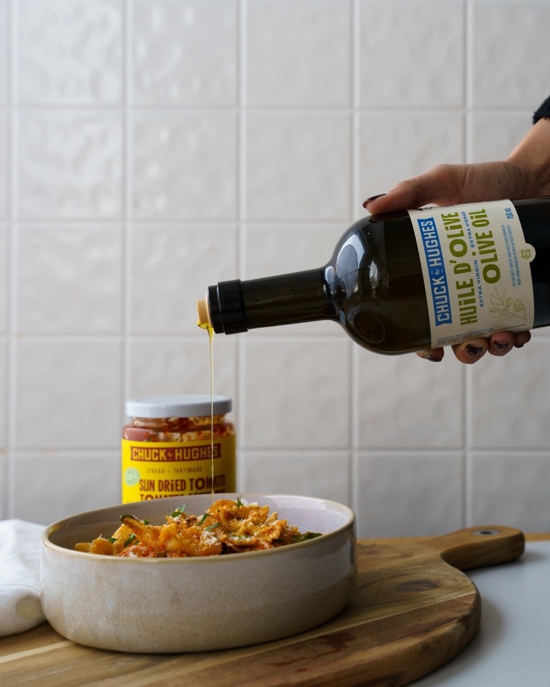 Une main verse l'huile d'olive Chuck Hughes sur un plat de pâtes, à côté d'un pot de tartinade de tomates séchées
